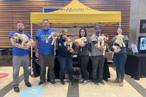 A wide angle photo of six people in a building, standing under a Manitoba Hydro branded tent, each smiling and holding a dog.