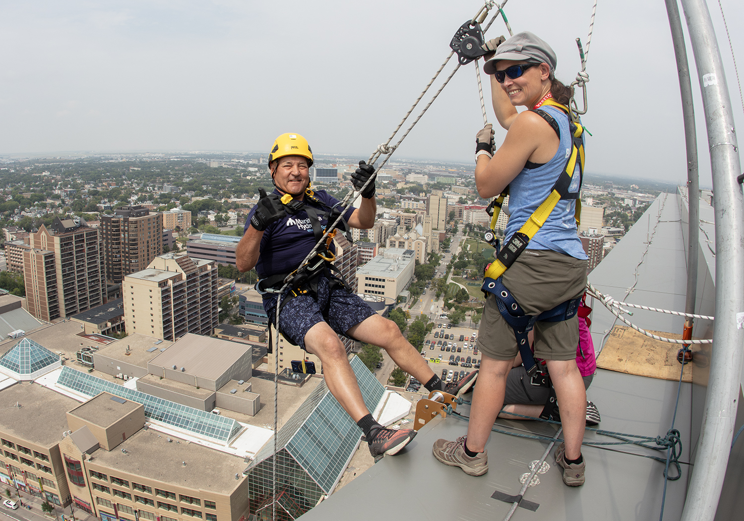 Two people stand at the top of Manitoba Hydro’s 23-story head office building before they rappel down the side of it in support of Manitobans with disabilities, their families, and their communities.