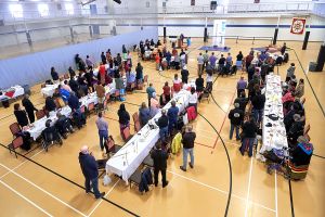 An overhead shot of a large group of people standing aside long banquet tables in a gymnasium. In the distance, a man and woman are at a podium speaking.